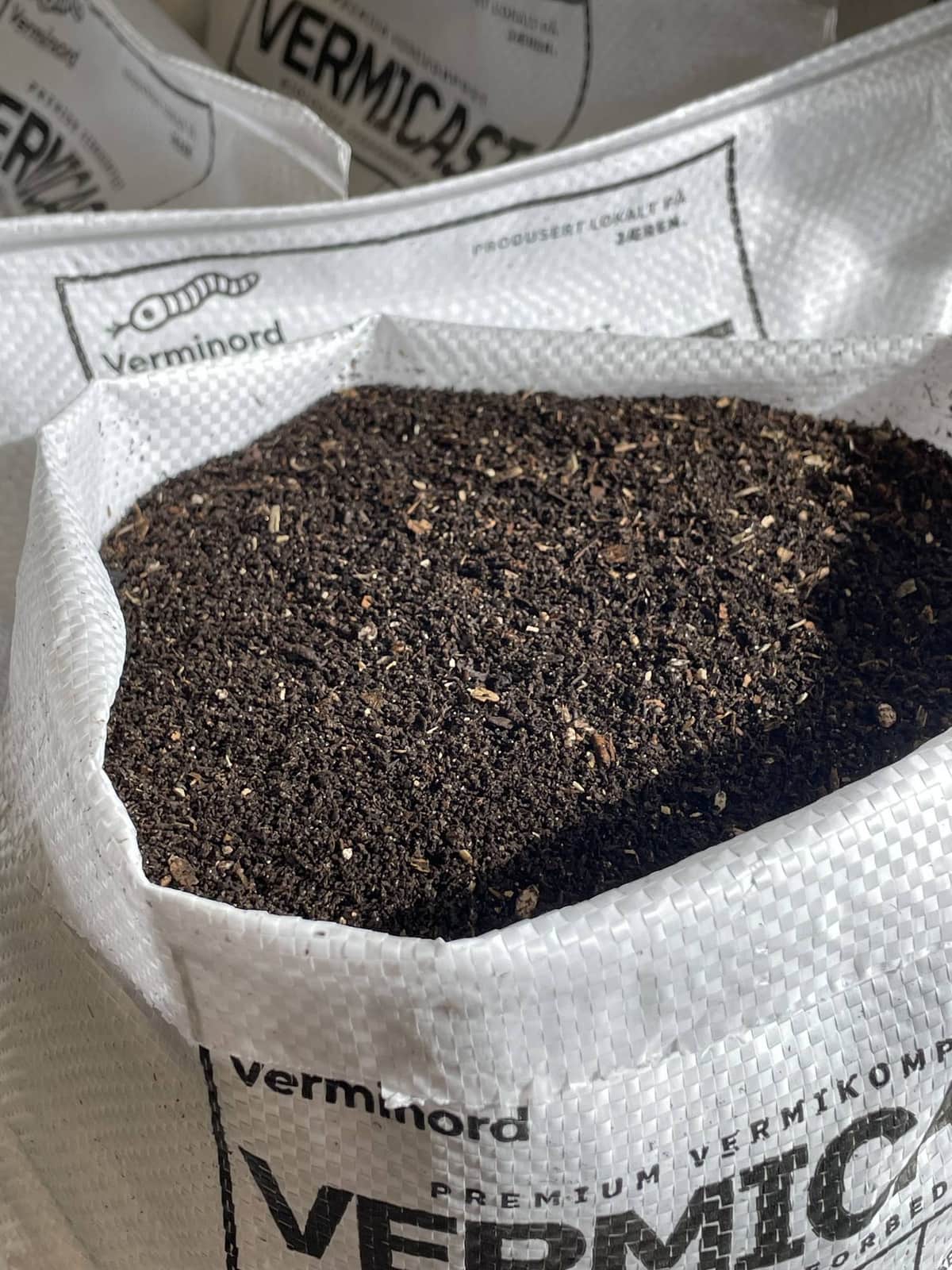 Åpen hvit vevd sekk merket VermiCast, fylt med mørk vermikompost. Flere lukkede sekker med Verminord-etikett står bak.